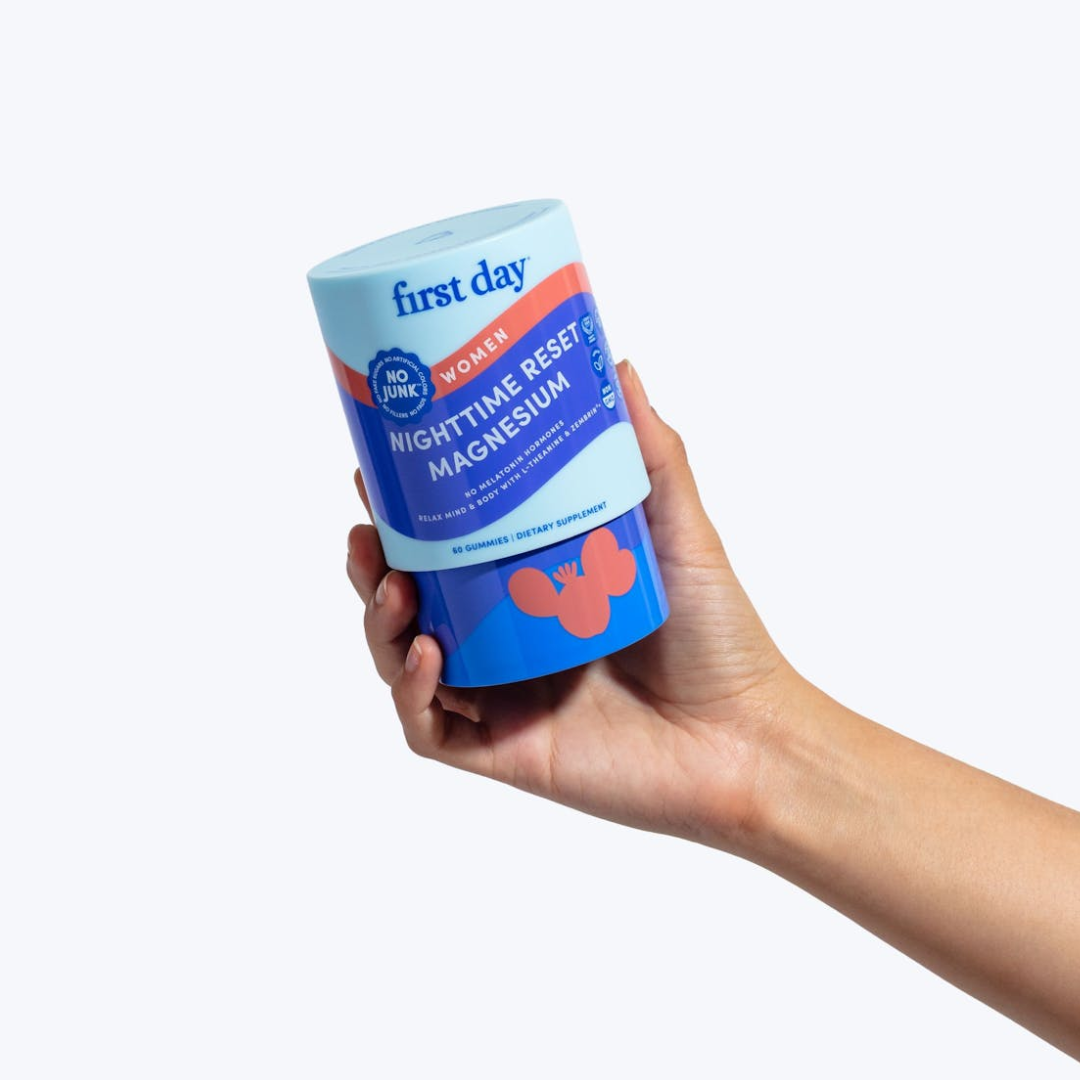 A hand holds a blue and purple container labeled first day Women Nighttime Reset Magnesium with an orange accent and white text on a plain light background.