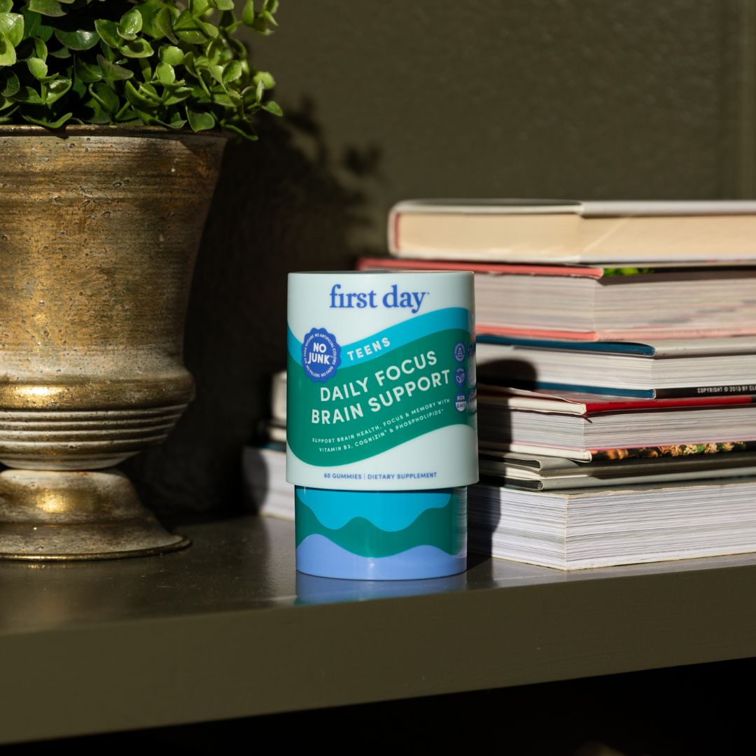 A container labeled first day Teens Daily Focus Brain Support sits on a shelf next to a stack of magazines and a potted plant. Sunlight partially illuminates the scene.