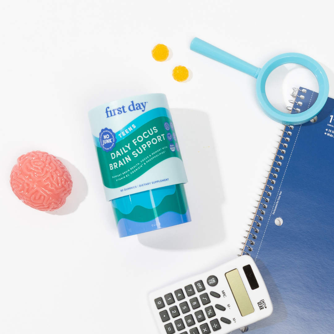 A container labeled first day Daily Focus Brain Support is surrounded by a toy brain, two yellow gummies, a magnifying glass, a spiral notebook, and a calculator on a white background.