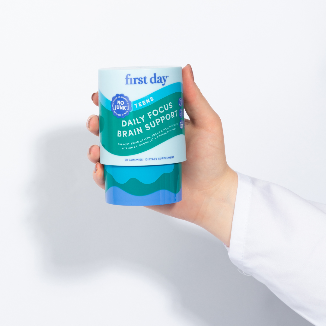 A hand in a white sleeve holds a container labeled “first day Teens Daily Focus Brain Support” with a turquoise and blue design. The container claims “No Junk!” and contains 60 gummies as a dietary supplement.