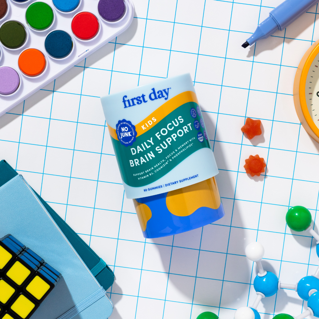 A bottle of First Day Kids Daily Focus Brain Support supplements is placed on a blue grid surface, surrounded by school supplies such as colored paints, a Rubiks cube, a clock, books, and science model pieces.