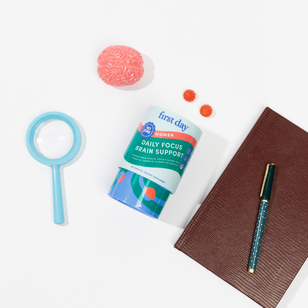 A canister labeled first day Women Daily Focus Brain Support sits on a white surface surrounded by a blue magnifying glass, a pink brain model, two red gummies, a teal pen, and a brown notebook.