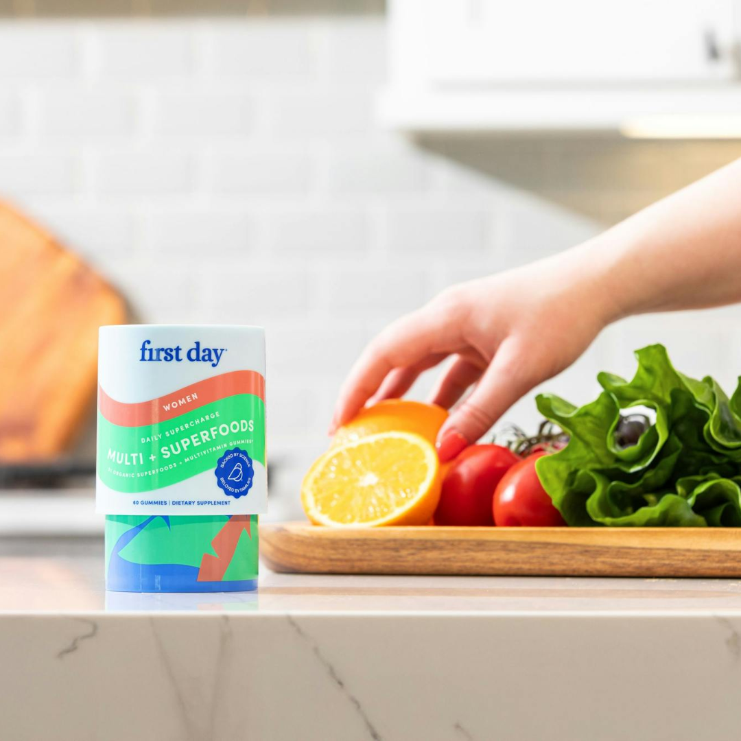 A hand holds half a lemon over a wooden cutting board with lettuce and tomatoes, next to a container labeled first day women’s multi + superfoods on a kitchen counter.