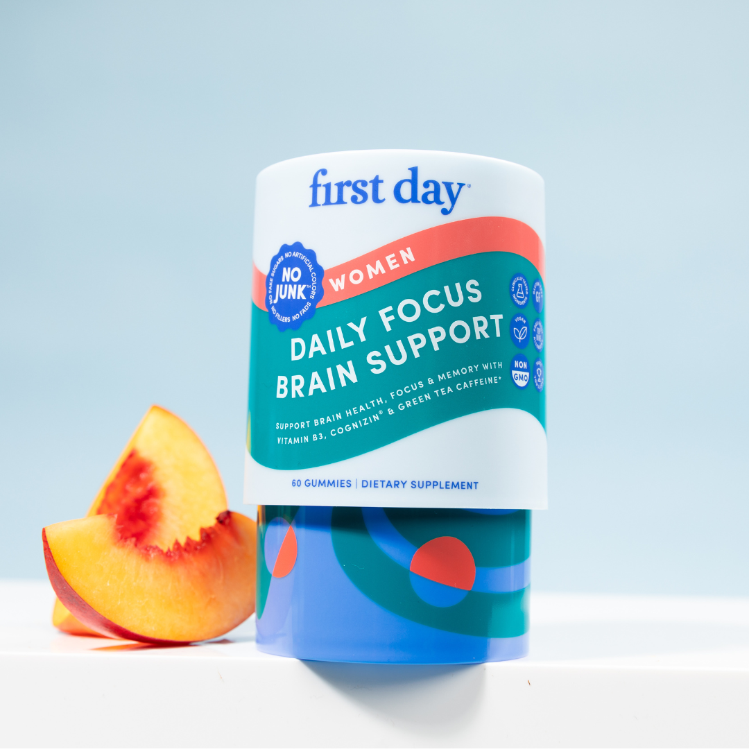 A container of First Day Women Daily Focus Brain Support gummies stands upright next to a peach slice on a white surface with a light blue background. The label highlights brain support and dietary supplement benefits.