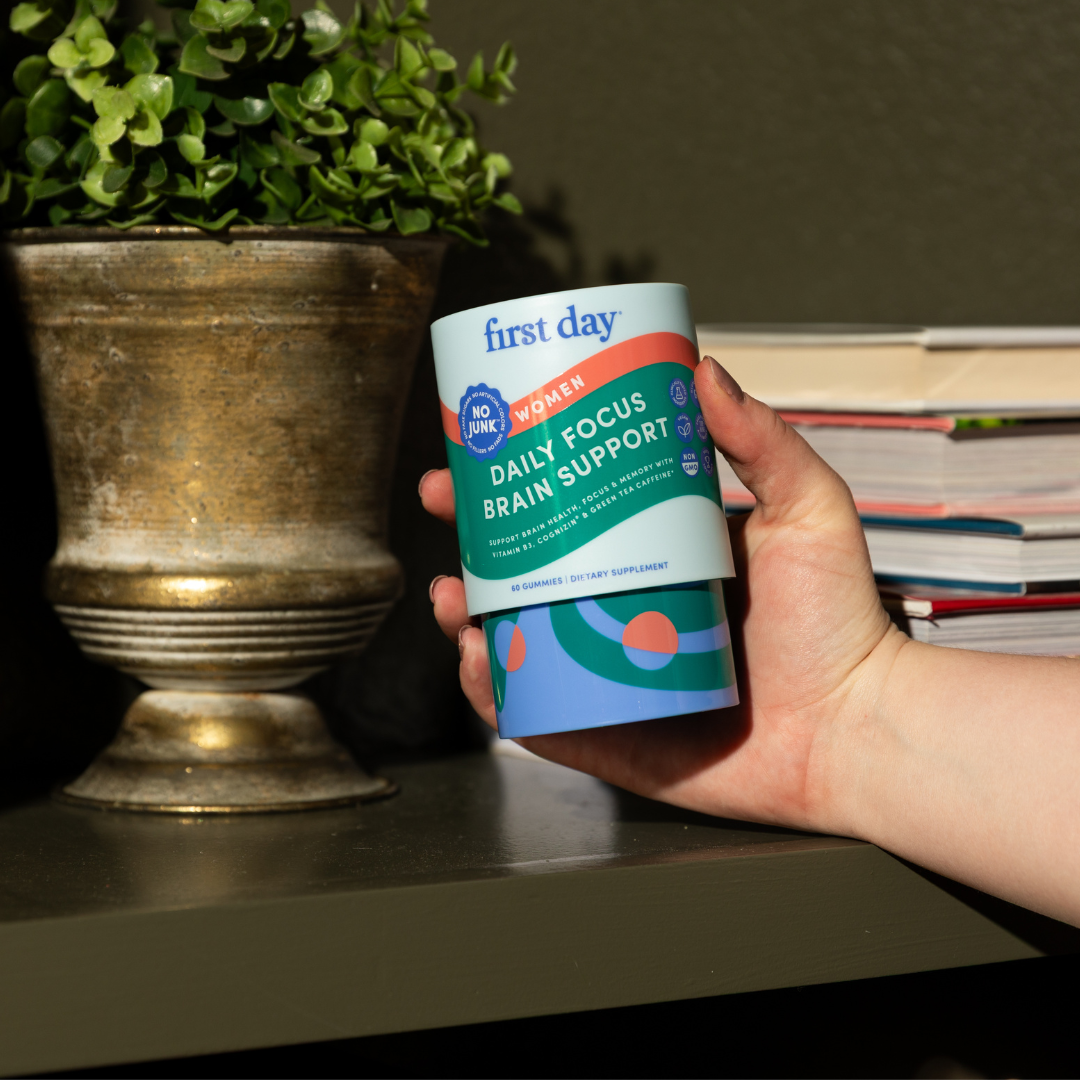 A hand holds a container labeled first day Daily Focus Brain Support for Women in front of a plant pot and a stack of books on a shelf. The container is colorful and features a NO JUNK badge.