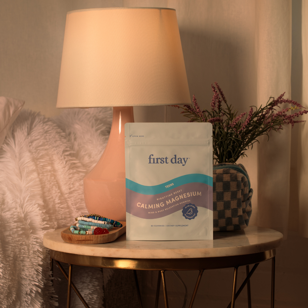 A bag of First Day Calming Magnesium dietary supplement sits on a marble bedside table next to a pink lamp, a potted plant, and a wooden tray with colorful beaded bracelets. Cozy bedroom decor is visible in the background.