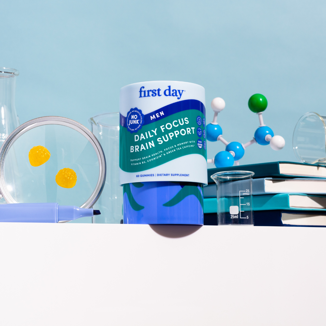 A container of First Day Men Daily Focus Brain Support gummies sits on a table with two yellow gummies, a pen, stacked books, molecular model, and glass labware against a light blue background.
