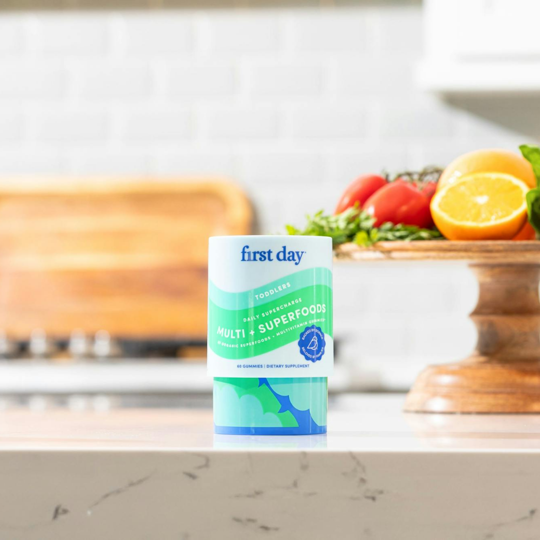 A container of First Day Toddler Multivitamin & Superfoods stands on a kitchen counter, with fresh vegetables and a halved lemon on a wooden tray in the background.
