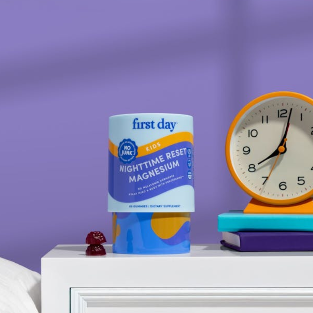 A container of First Day Kids Nighttime Reset Magnesium gummies sits on a white nightstand beside stacked books and an analog clock against a purple background.