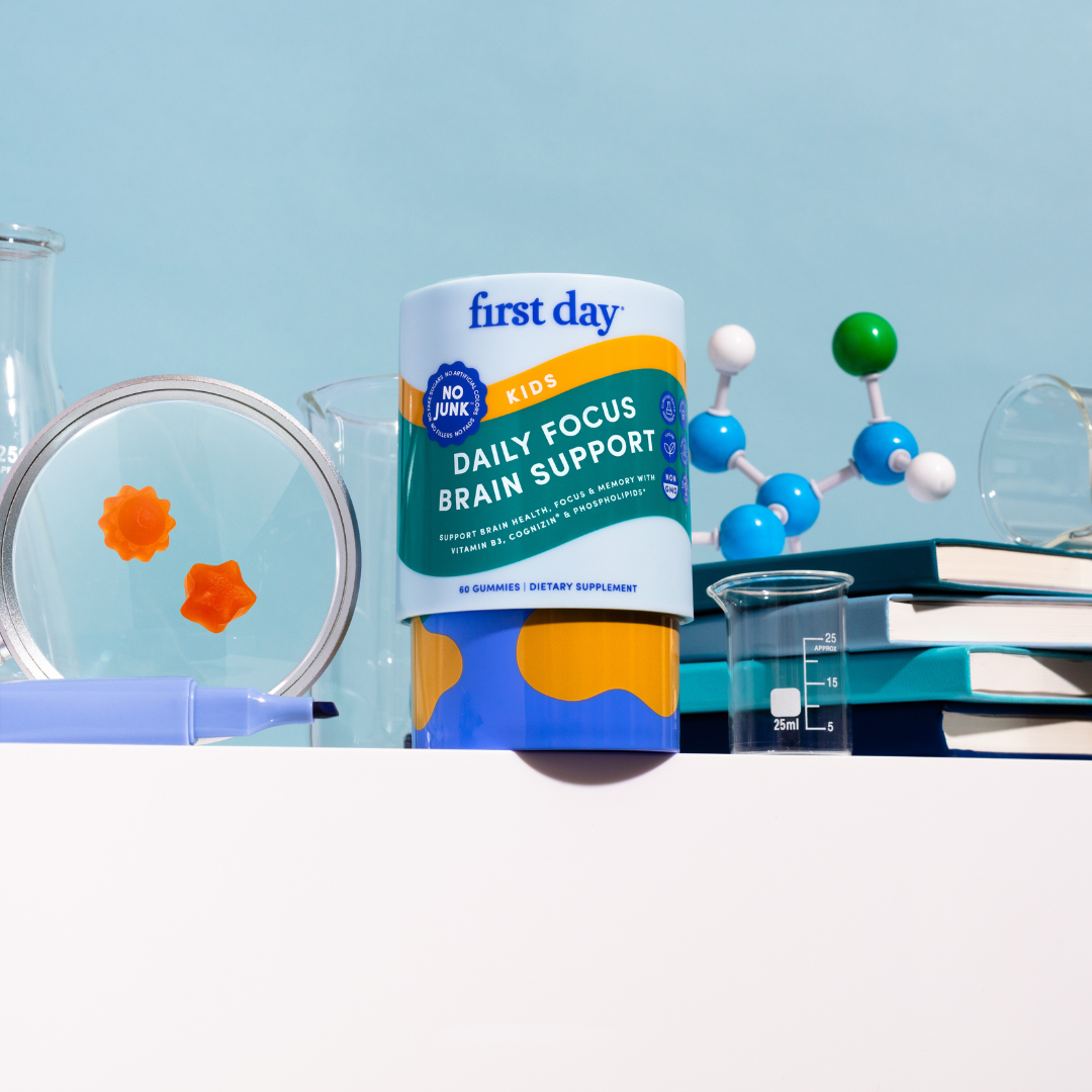 A container of First Day Kids Daily Focus Brain Support gummies is on a white surface, surrounded by scientific lab equipment, books, a pen, and a molecular model, with an orange gummy visible in a petri dish.
