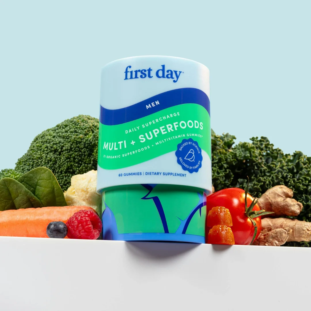 A container of First Day Men’s Multi + Superfoods gummies is displayed in front of fresh vegetables, including broccoli, kale, carrot, tomato, and apple, against a light blue background.