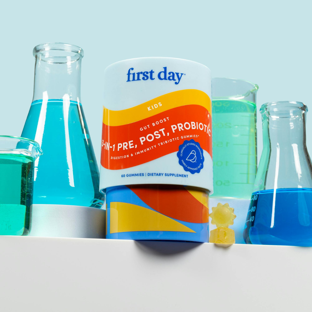 A container of First Day Kids 3-in-1 Pre, Post, Probiotic gummies sits on a shelf surrounded by laboratory glassware filled with blue and green liquid, with two yellow gummies placed in front of the container.