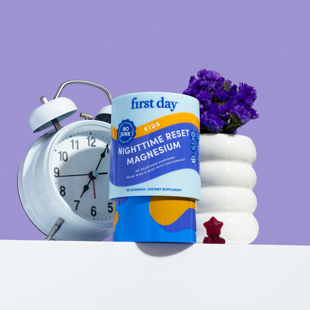 A container of First Day Kids Nighttime Reset Magnesium gummies is displayed next to a light blue alarm clock, a small red star-shaped object, and a white vase with purple flowers against a purple background.
