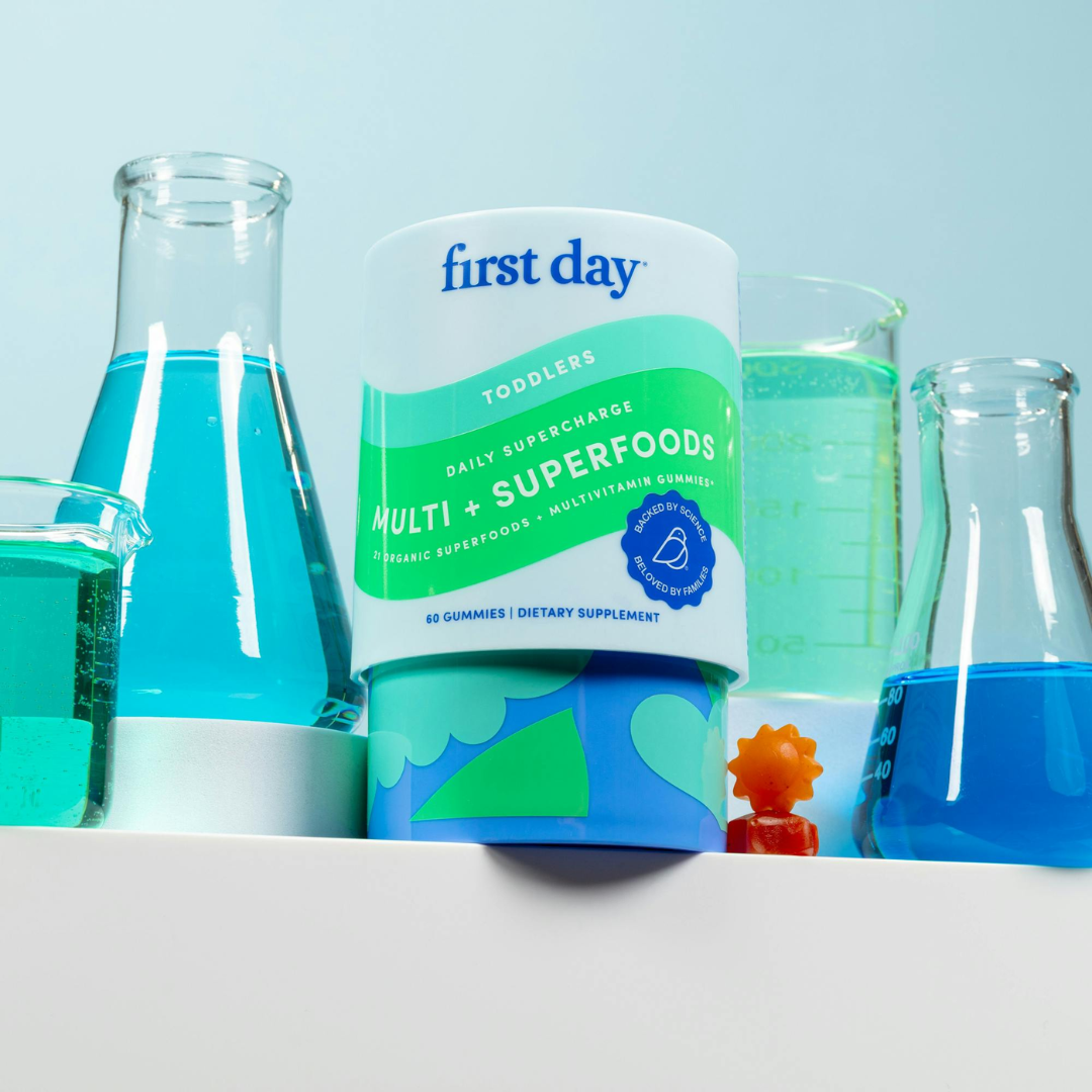 A container of First Day Toddlers Multi + Superfoods gummy vitamins is displayed in front of laboratory beakers filled with blue liquid, with a small orange figurine beside the container.