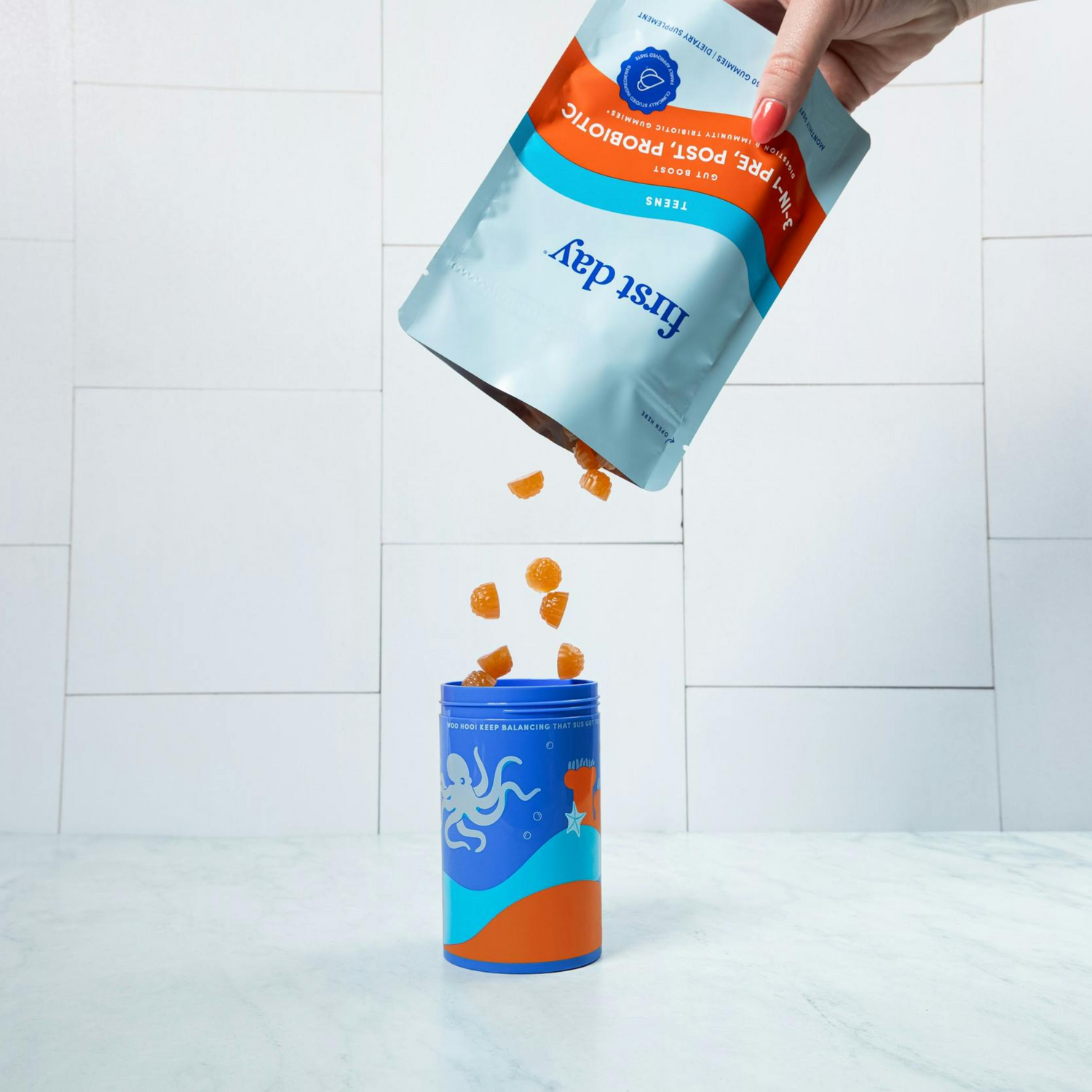 A hand pours orange gummy supplements from a resealable pouch labeled first day Fiber + Post Probiotic into a blue container with an octopus illustration, set on a white marble surface with a tiled background.