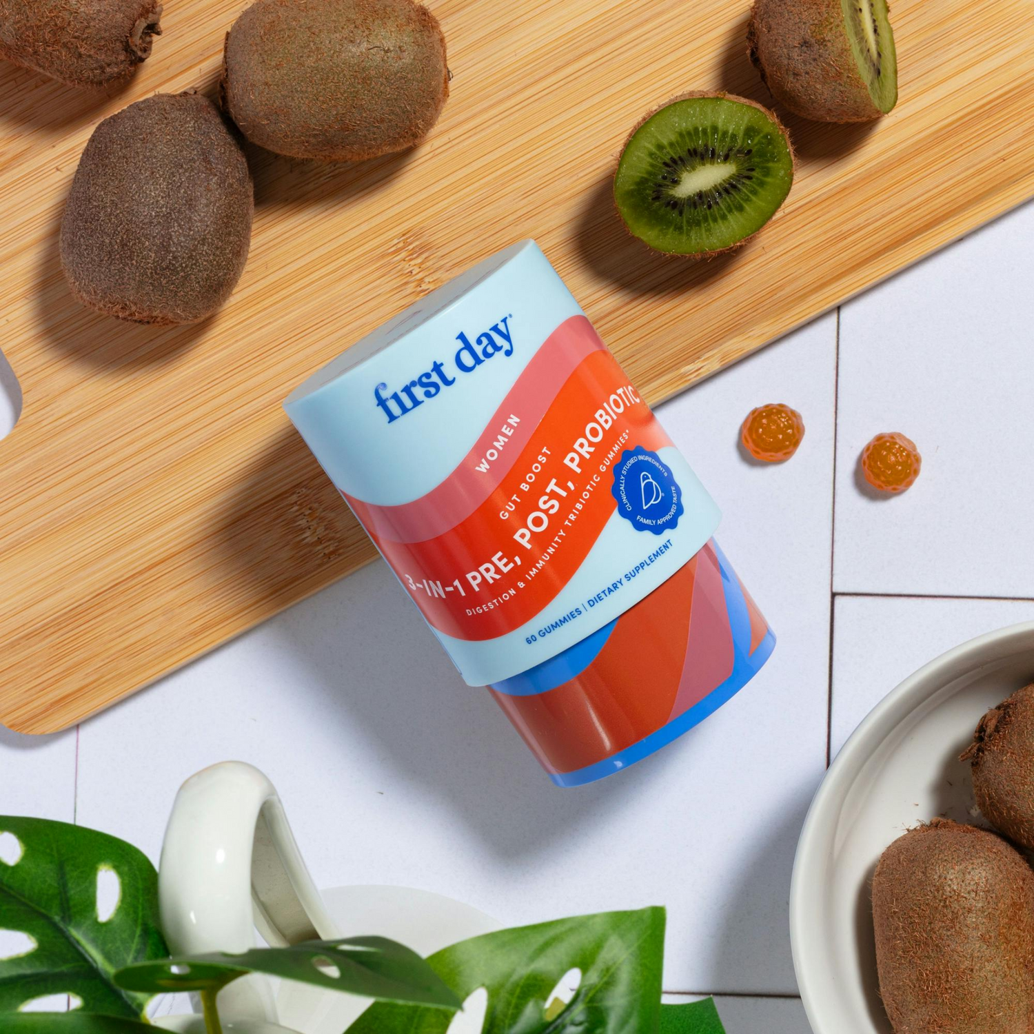A container of First Day Women’s Probiotics lies on a wooden board surrounded by whole and sliced kiwis, two gummy supplements, and a leafy plant, all on a white tile surface.