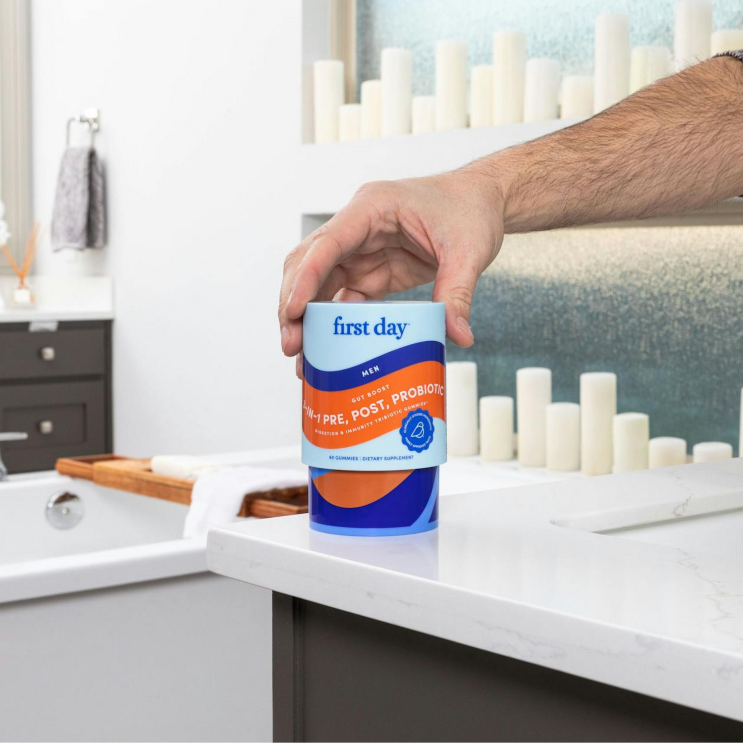 A person’s hand is holding a container labeled “first day Kids Multi + Pre, Post, Probiotics” in a bathroom with a white countertop, a bathtub, and neatly arranged candles in the background.