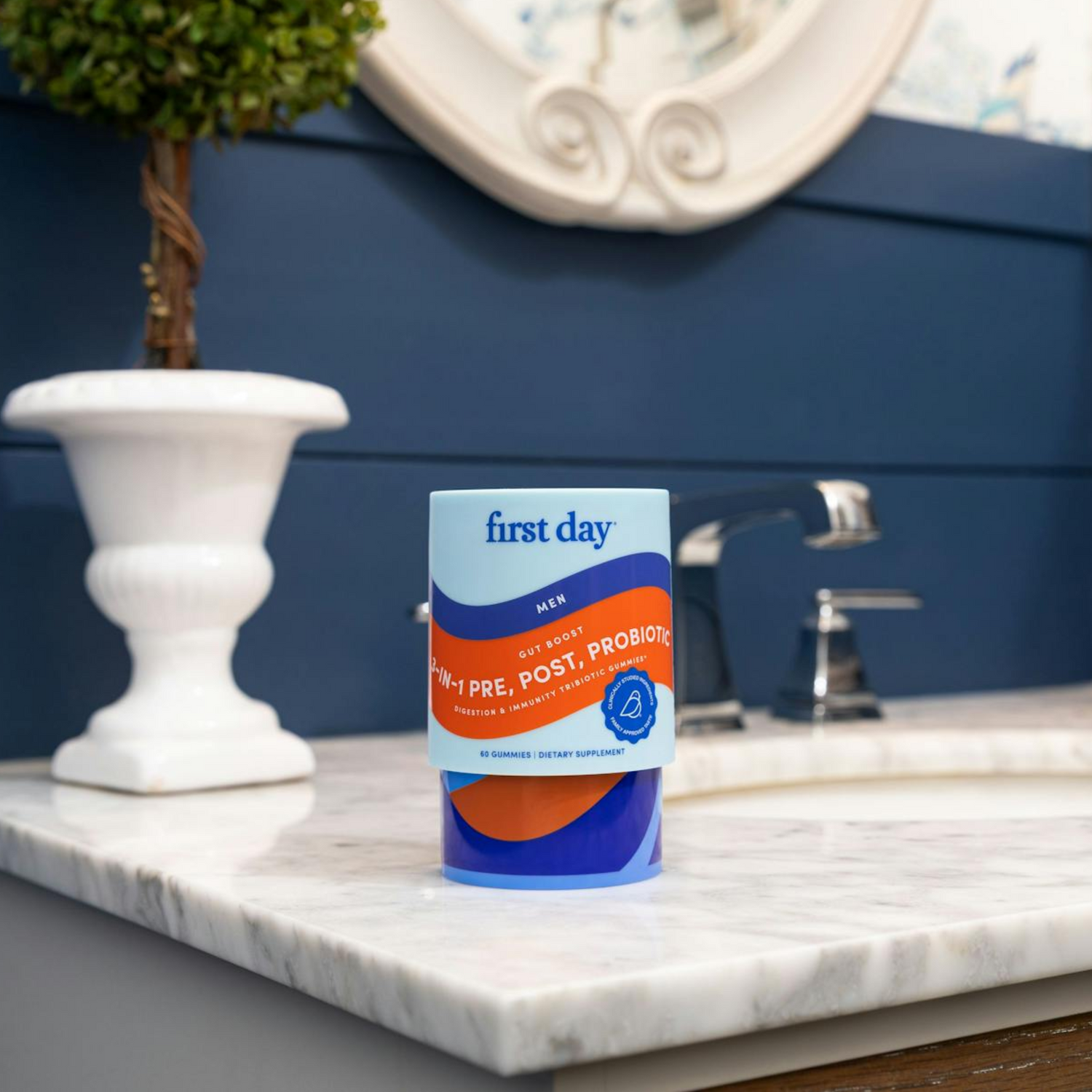 A container of first day mens gut health pre, post, and probiotic supplement sits on a marble bathroom countertop, with a white vase and plant in the background.