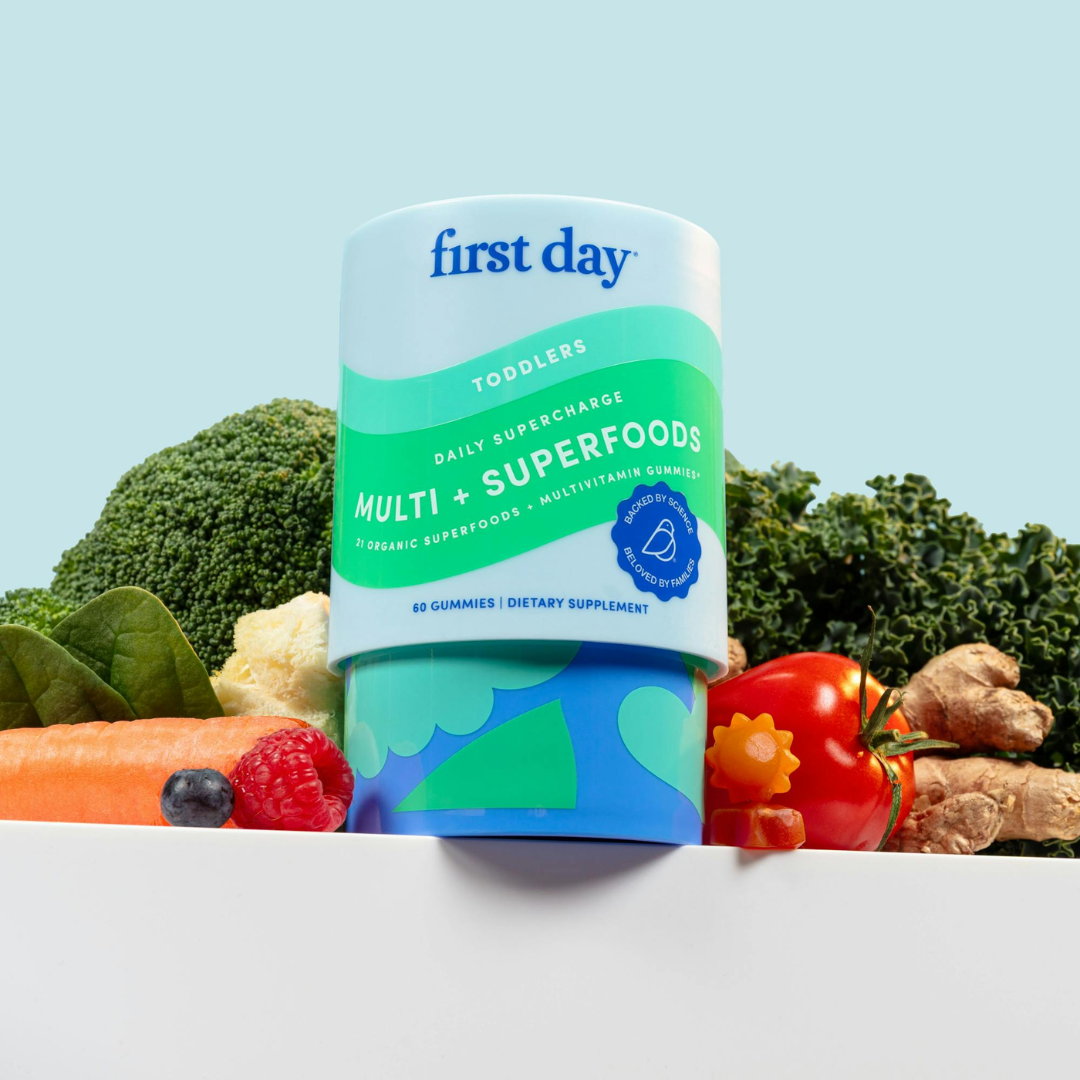 A container of First Day Toddlers Multi + Superfoods gummies is displayed among fresh vegetables and fruit, including broccoli, kale, carrot, tomato, and berries, against a light blue background.