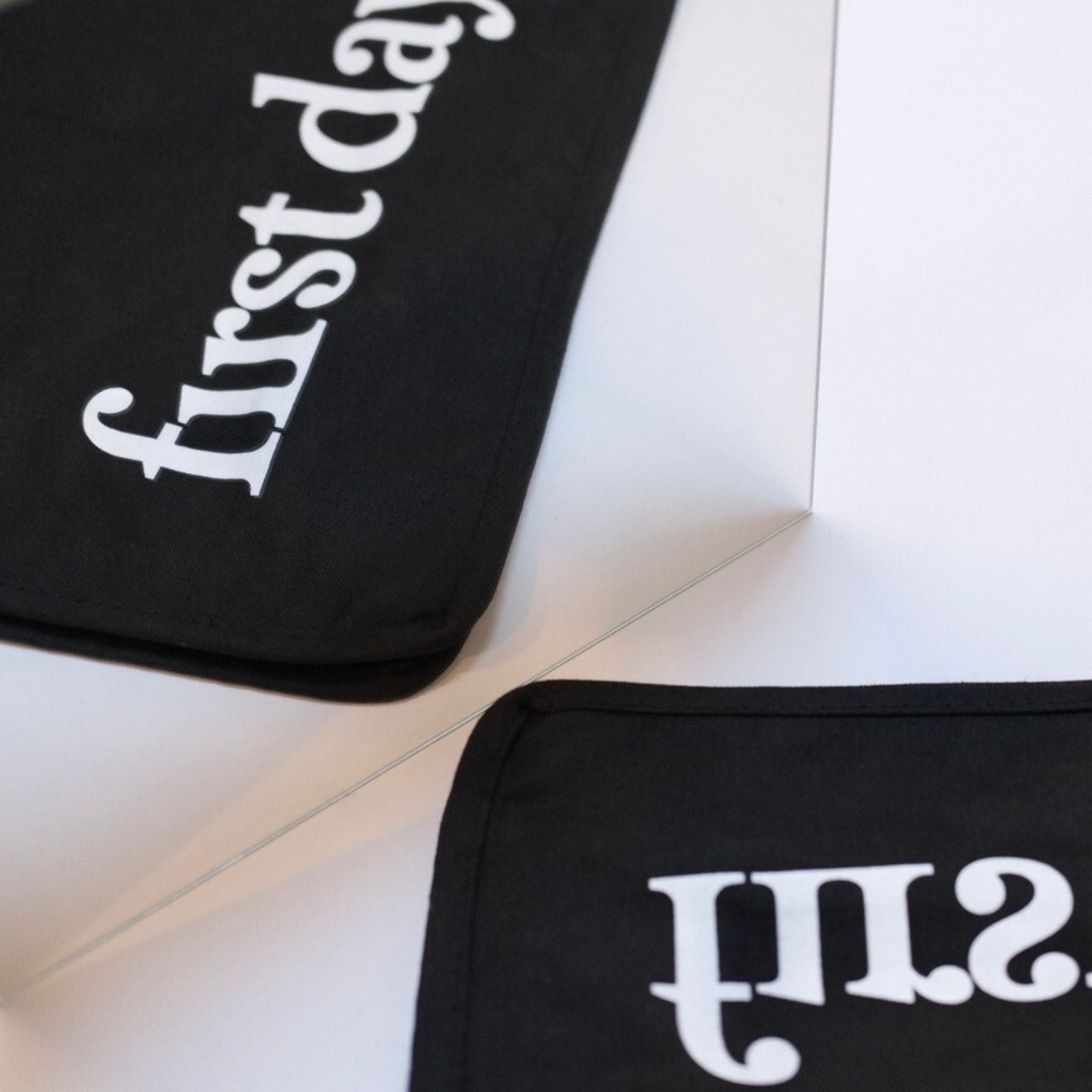 The Reflection Tote features “first day” in white, partially mirrored for a subtle hidden message. This black tote’s angled design on a white surface adds intrigue—ideal for anyone seeking a unique and stylish accessory.