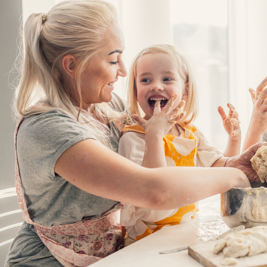 A smiling woman and a young girl with messy hands laugh together while baking in a sunlit kitchen, both wearing aprons and surrounded by dough on the table.