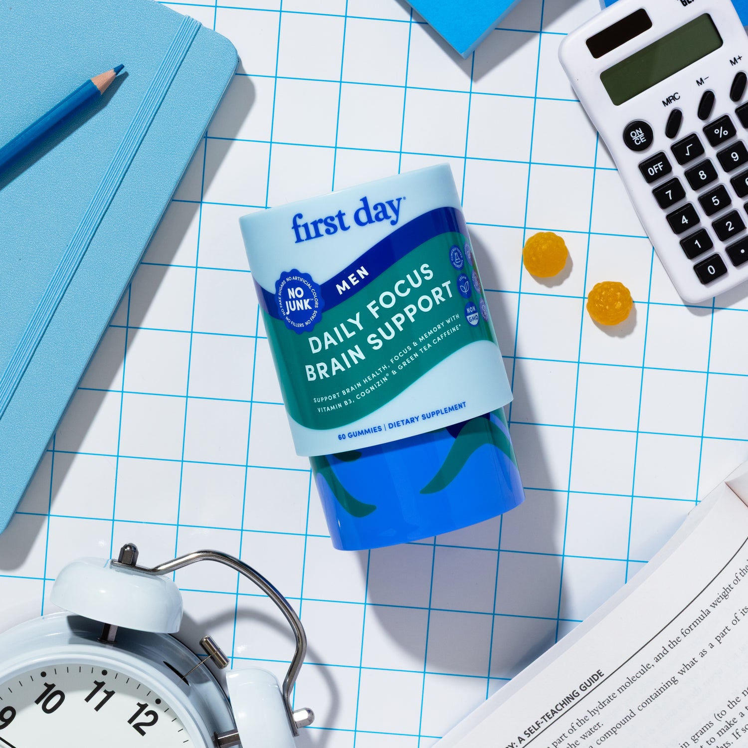 A container of First Day Daily Focus Brain Support supplements lies on a blue grid surface, surrounded by a white alarm clock, calculator, notebook, pen, two yellow gummies, and an open book.