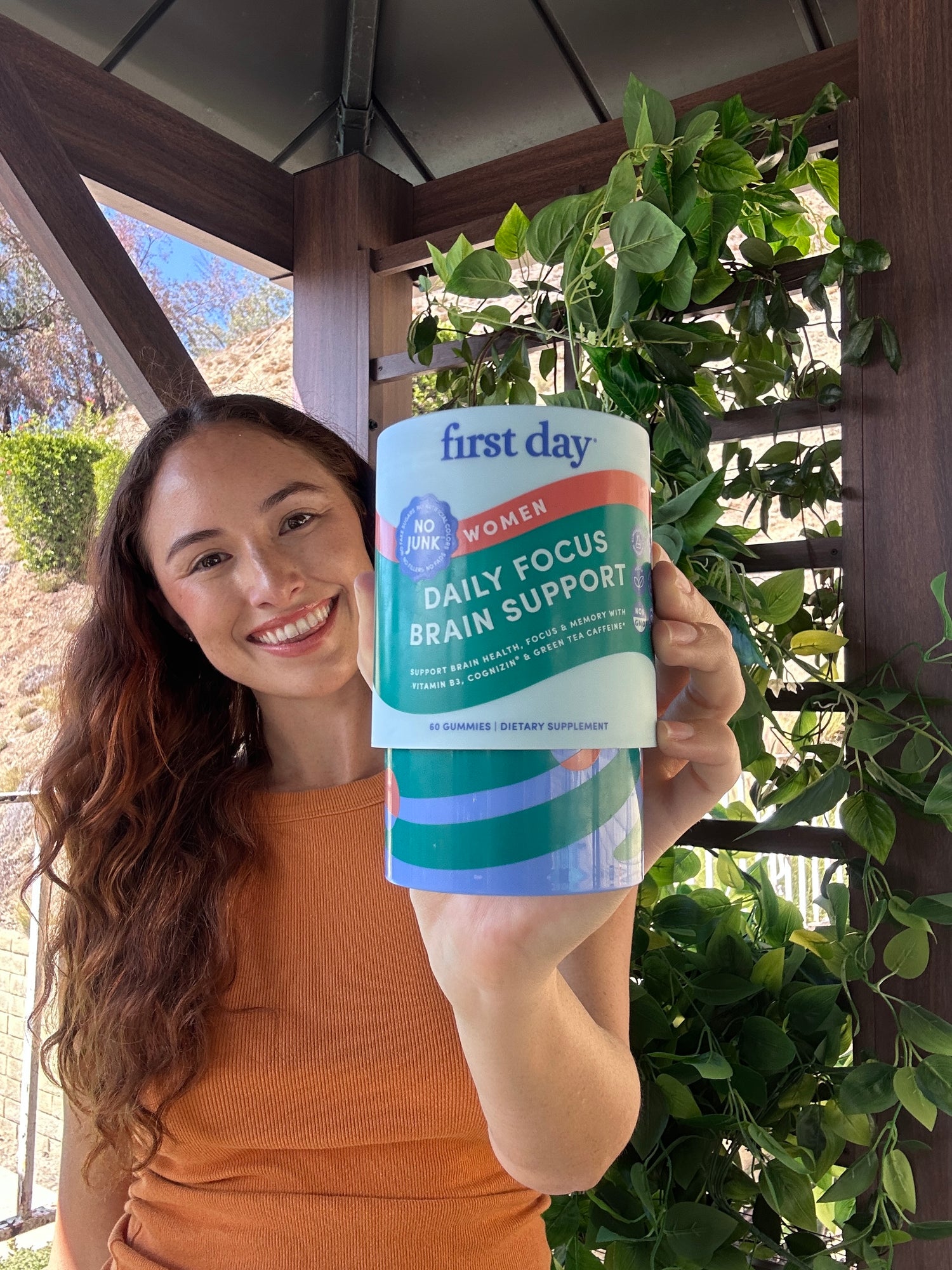 A smiling woman in an orange shirt holds up a large container of First Day Daily Focus Brain Support gummies for women. She stands outdoors in front of leafy green plants and a wooden structure.