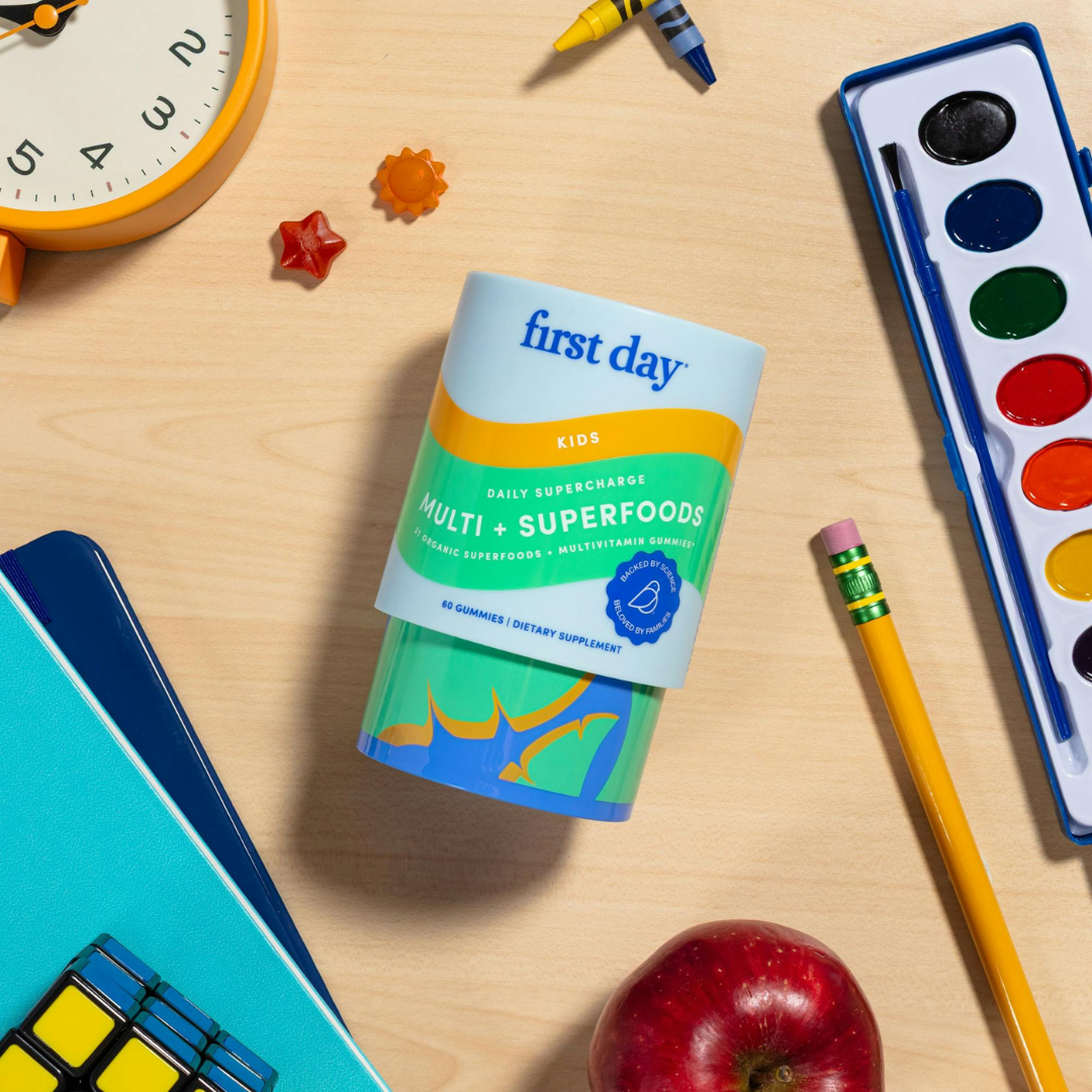A container of First Day Kids Multi + Superfoods gummy vitamins is on a wooden desk surrounded by a clock, notebook, pencil, watercolor paint set, apple, and small toy shapes.