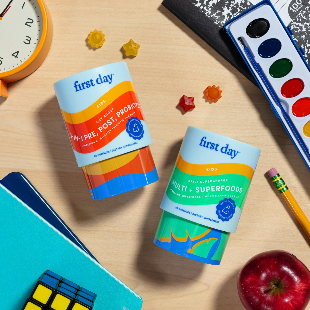 Two colorful bottles of First Day kids’ supplements are on a desk with a Rubik’s cube, pencils, a red apple, books, a clock, and watercolor paints, surrounded by small star-shaped objects.