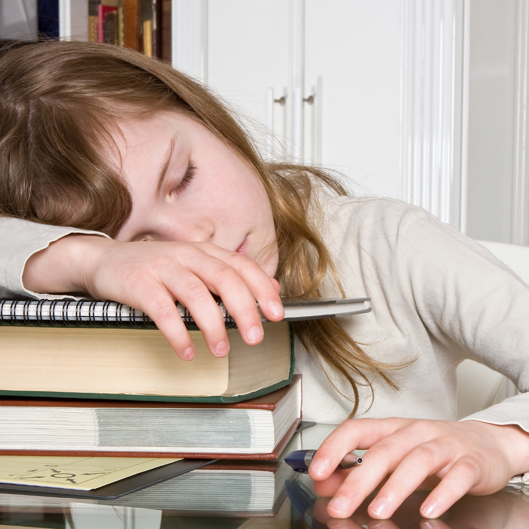 A young girl with light brown hair is asleep at a table, resting her head on a stack of books and notebooks, with her hand holding a pen. Papers are scattered nearby.