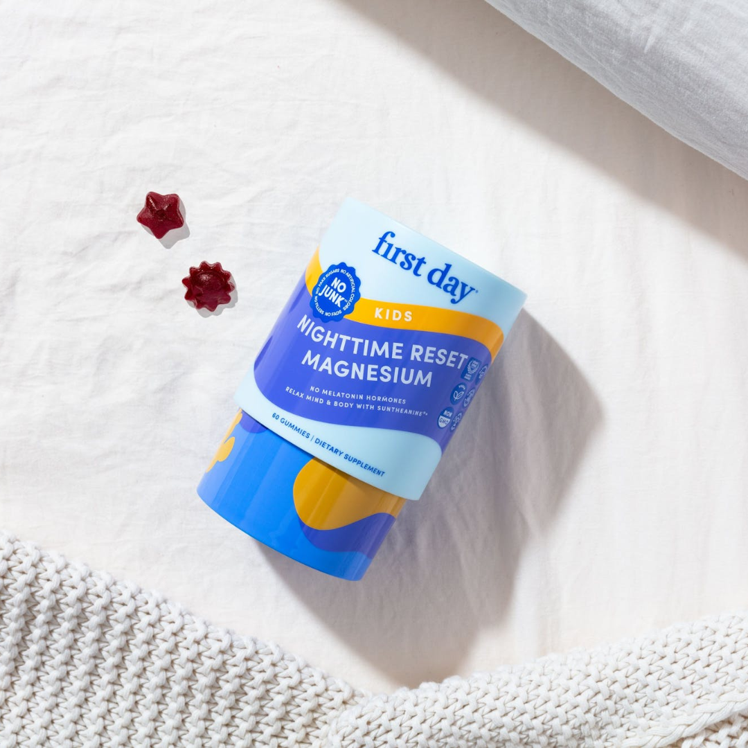 A container of First Day Kids Nighttime Reset Magnesium gummies lies on a white bed with a knitted blanket, pillow, and two red bear-shaped gummies beside the container.