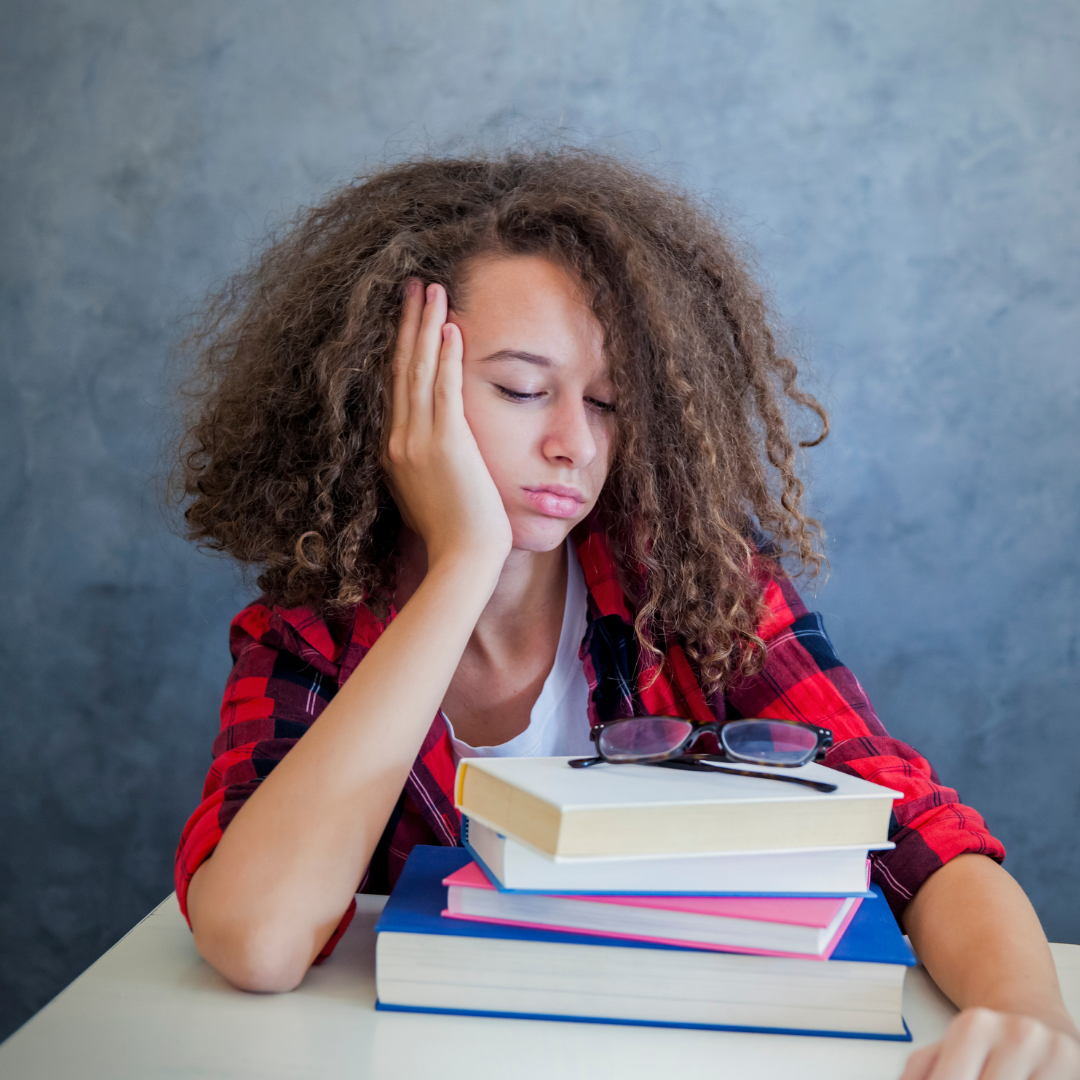 A tired student with curly hair, wearing a red plaid shirt, rests their head on one hand and looks down at a stack of books and glasses on a desk.
