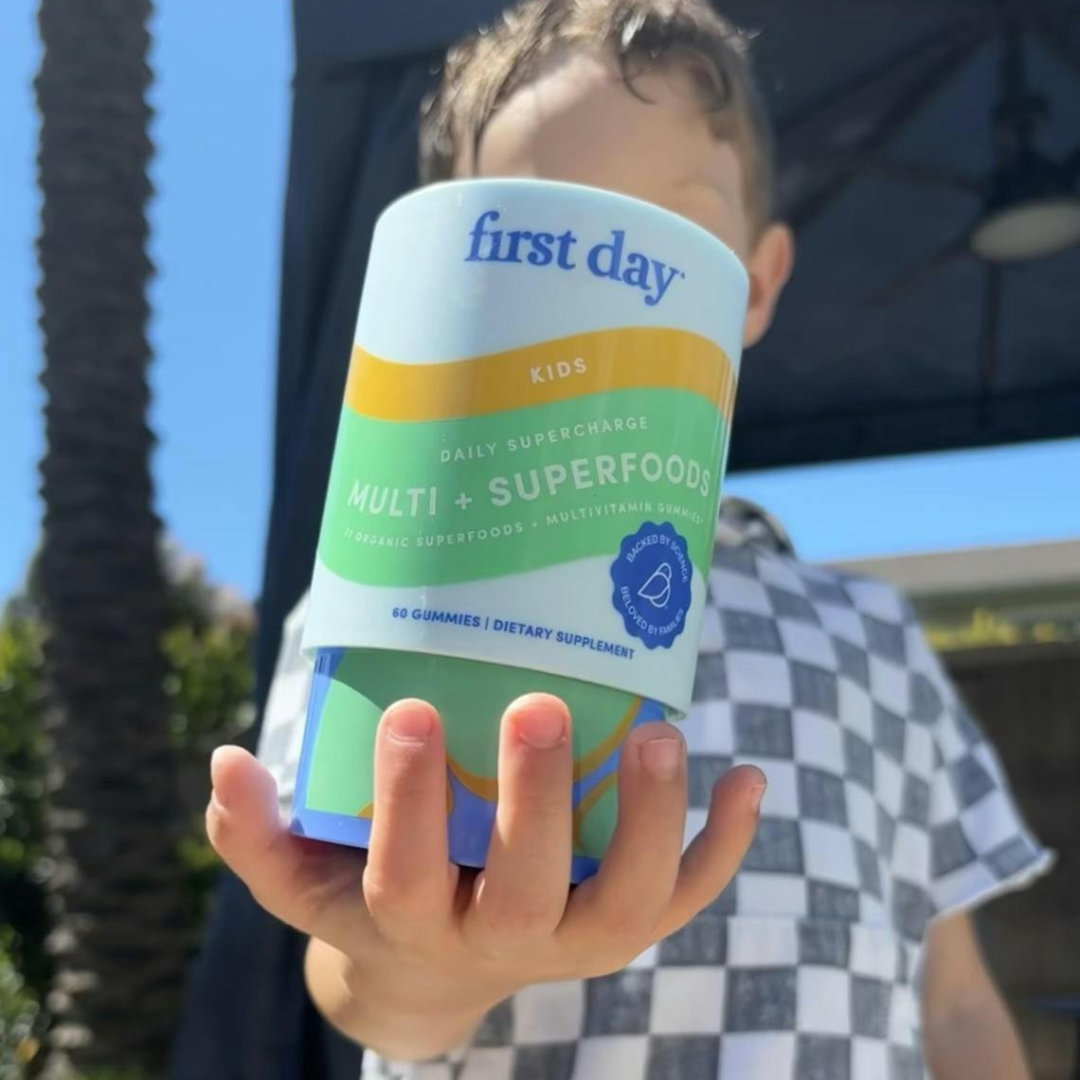 A child in a checkered shirt holds a container of First Day Kids Multi + Superfoods gummies toward the camera, with an outdoor background of blue sky, trees, and a shaded structure.