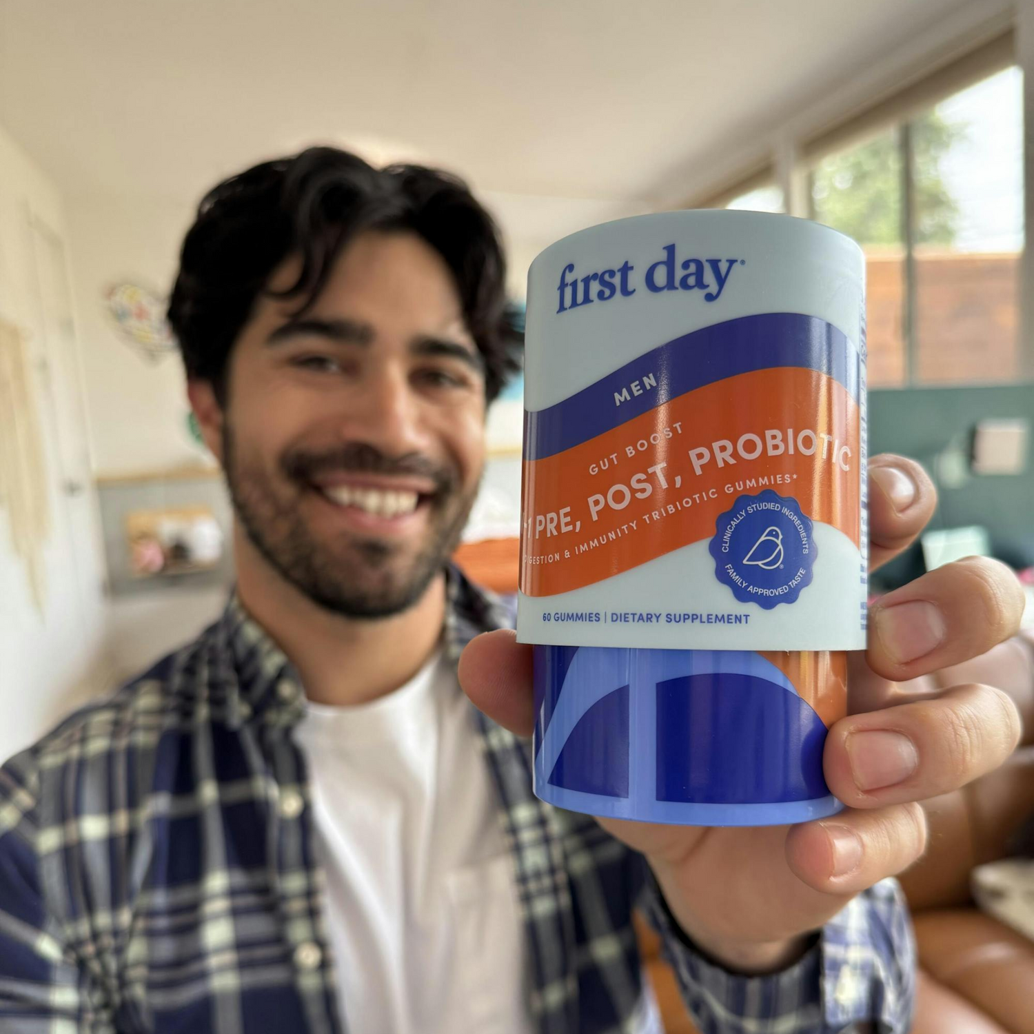 A smiling man holds up a container of first day mens pre, post, and probiotic dietary supplement gummies, with the product label clearly visible in focus.