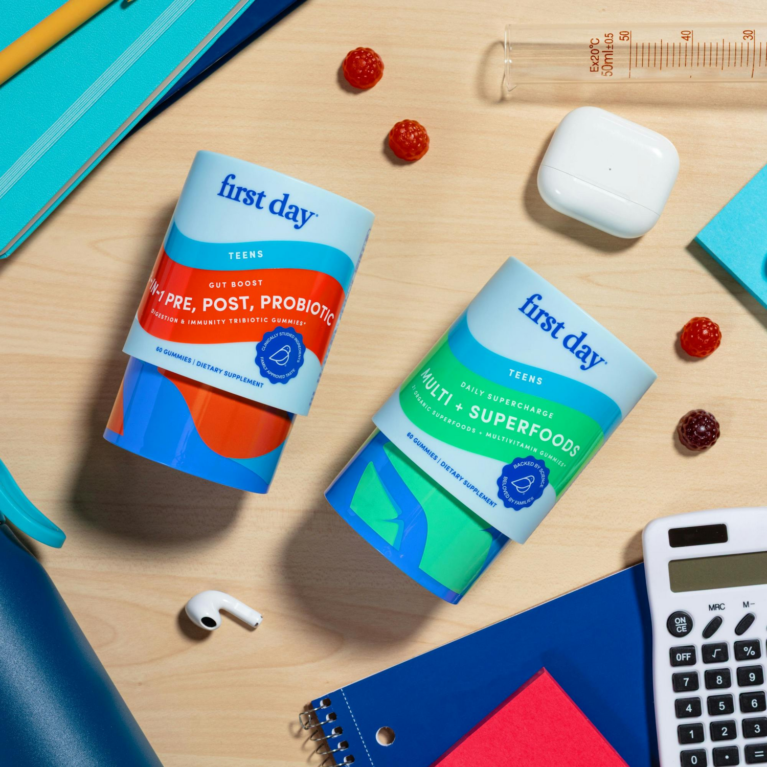 Two first day supplement containers for teens are placed on a desk with AirPods, a calculator, a notebook, a beaker, scattered gummies, and office supplies surrounding them.