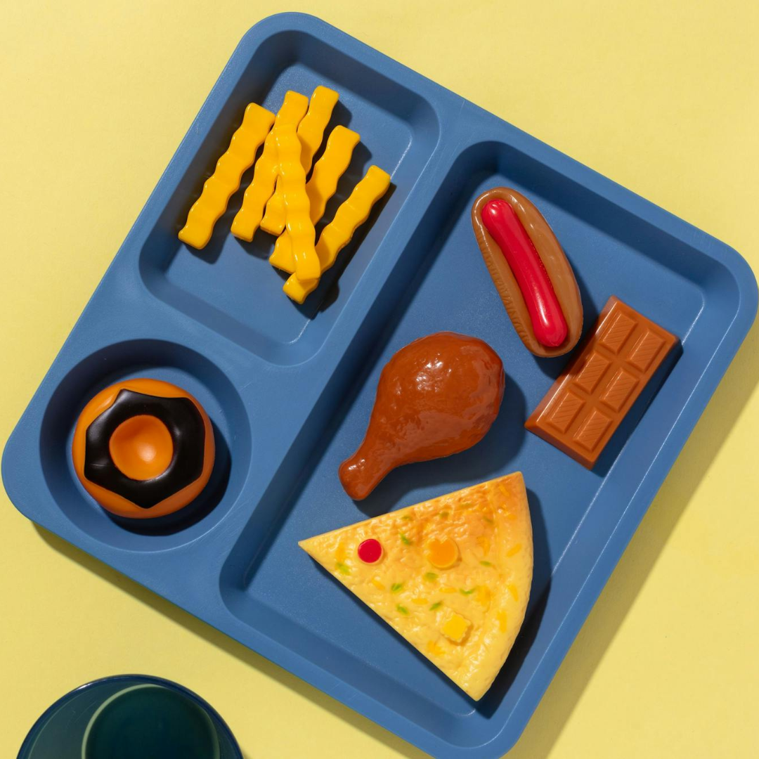 A blue lunch tray with toy food items, including crinkle-cut fries, a hot dog, a chicken drumstick, a slice of pizza, a chocolate bar, and a cup with a black and orange dessert, on a yellow background.