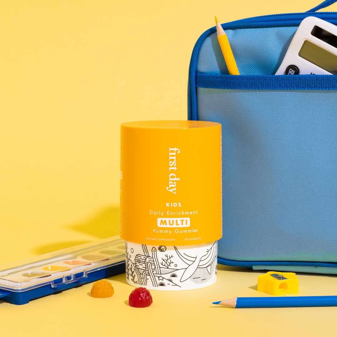 A yellow container of First Day Kids Daily Enrichment Multi Gummies stands next to a blue school bag, pencil, calculator, watercolor set, sharpener, and two gummy vitamins, all set against a yellow background.