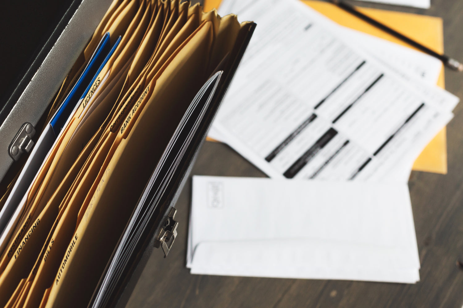 A close-up of a briefcase filled with organized file folders, with documents, envelopes, and a pencil scattered on a wooden desk in the background.