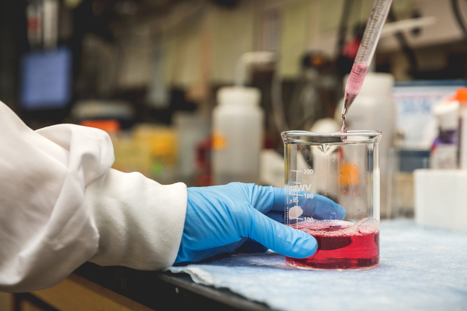 A person wearing a blue glove holds a beaker with pink liquid, while another hand uses a pipette to add more liquid, in a laboratory setting with blurred equipment in the background.
