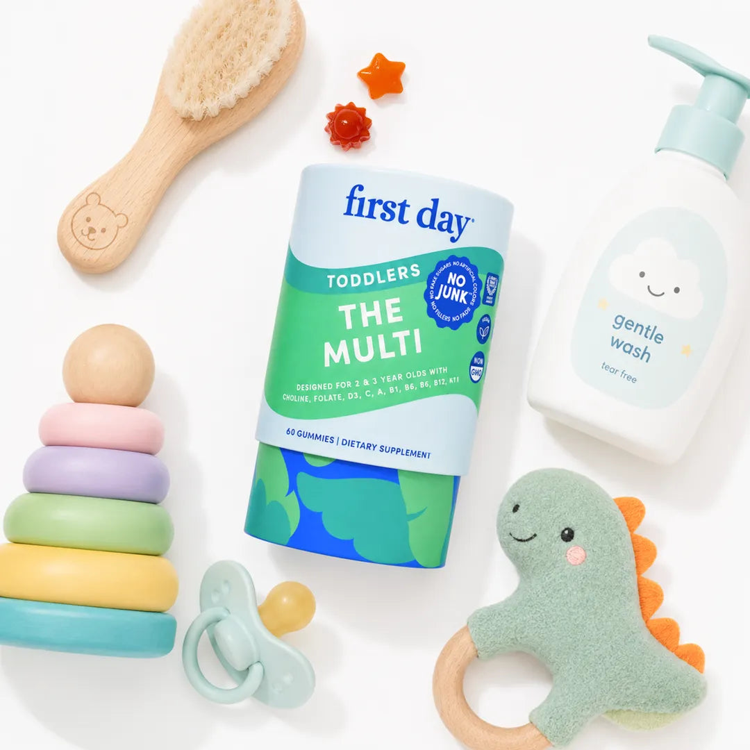 A styled flat-lay of baby and toddler products on a white background, featuring a container of First Day Toddlers &
