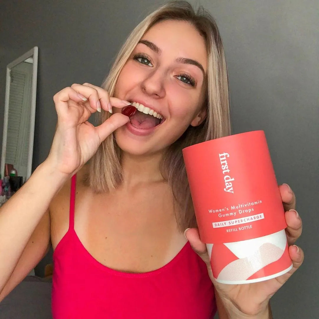 A smiling woman in a pink top holds a container labeled first day women’s multivitamin gummy drops in one hand and is about to eat a gummy drop with the other hand.