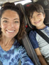 A woman and a young boy sit side by side in a car, both smiling at the camera. The boy is in a car seat, wearing a seatbelt and a blue shirt; the woman is wearing a blue tie-dye top.