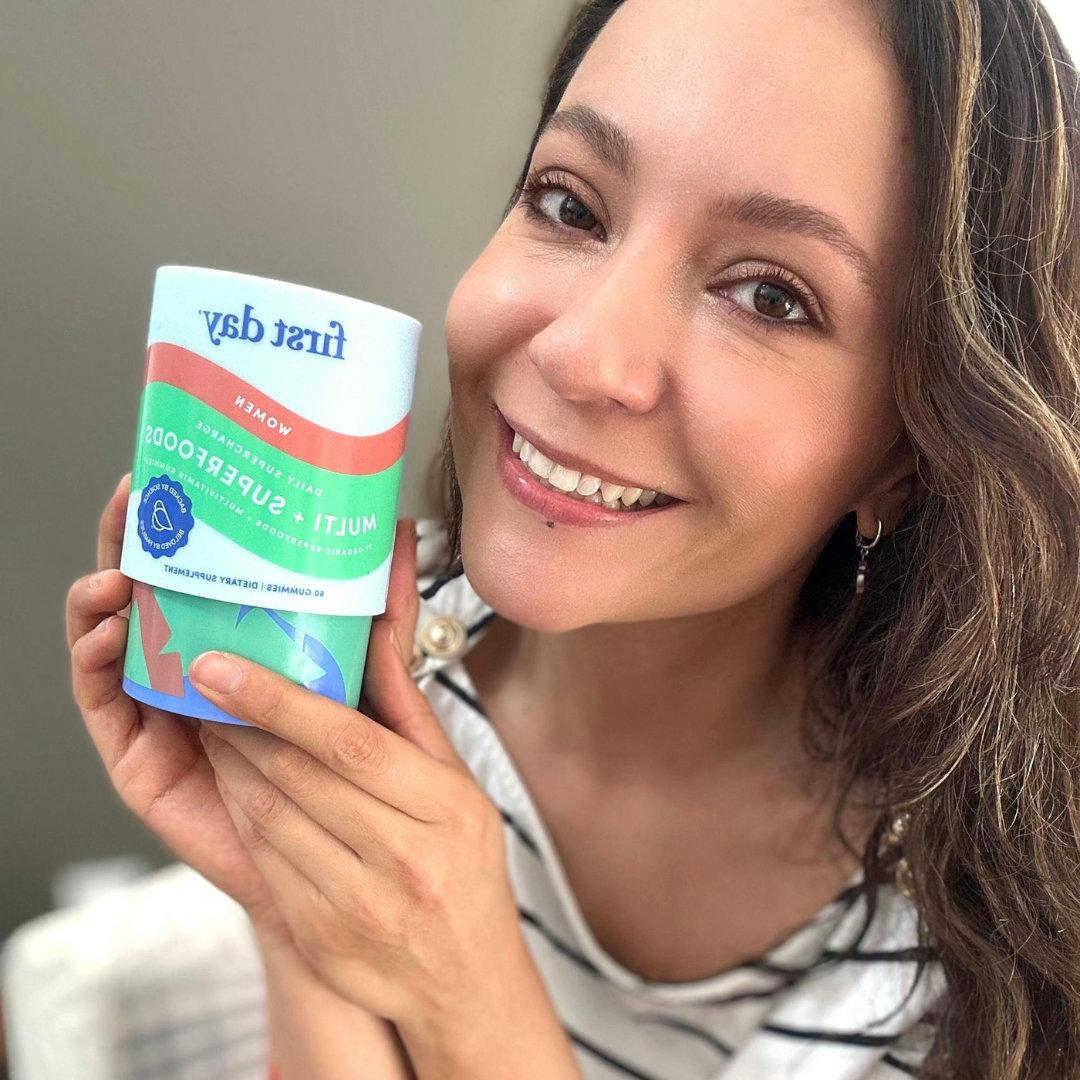 A smiling woman with long brown hair holds up a container of First Day Women’s Multivitamin, showing the label to the camera. She is indoors and wearing a striped shirt.
