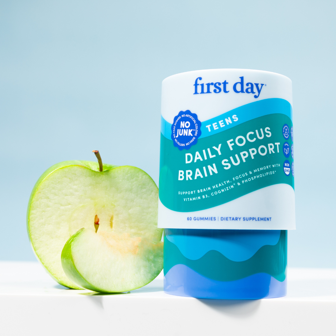 A container of First Day Teens Daily Focus Brain Support gummies is displayed next to a sliced green apple, with a light blue background. The label highlights support for brain health and memory.