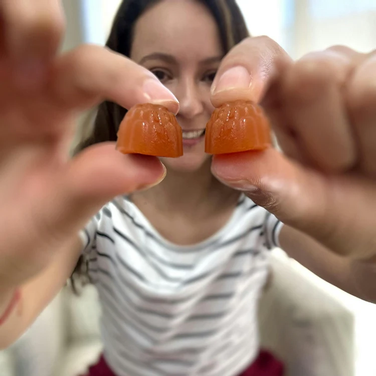 A woman in a striped shirt smiles and holds two orange gummy supplements close to the camera, one in each hand, showing them side by side.