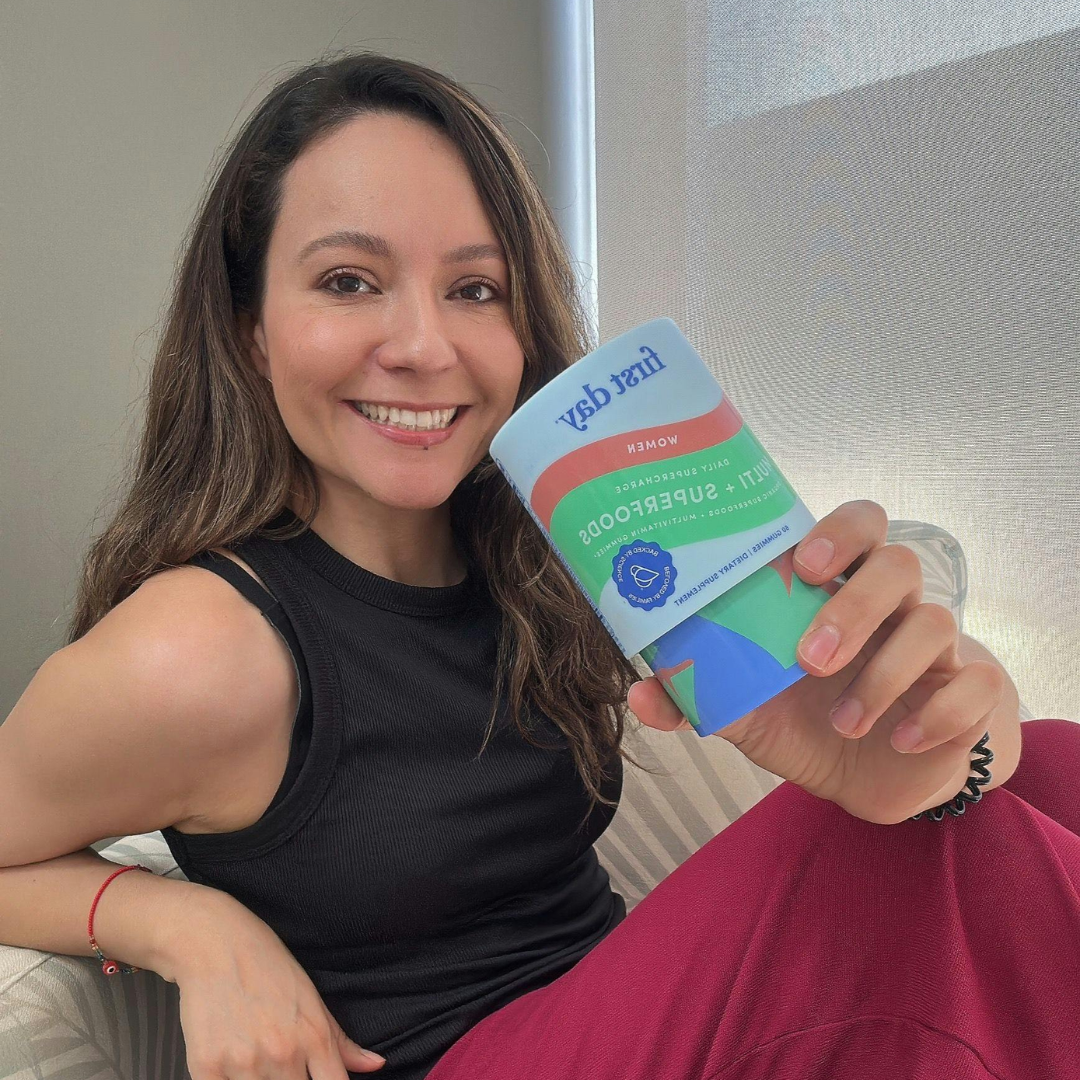 A smiling woman with long brown hair, wearing a black sleeveless top and red pants, holds up a package labeled 1st day women multivitamin superfood. She is sitting indoors near a window.