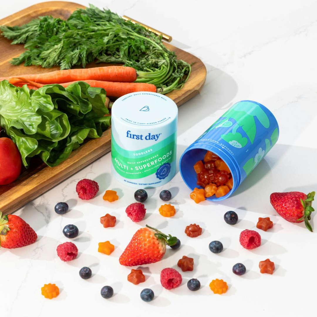Bottles of First Day Multi-Superfoods gummies are surrounded by scattered gummies, strawberries, blueberries, raspberries, and a cutting board with lettuce, carrots, and a tomato.