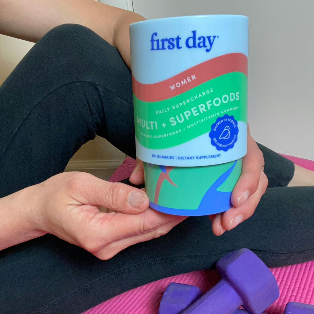 A person sitting on a pink yoga mat holds a container labeled first day WOMEN MULTI + SUPERFOODS dietary supplement gummies. Purple dumbbells rest nearby. The person is dressed in black workout clothes.