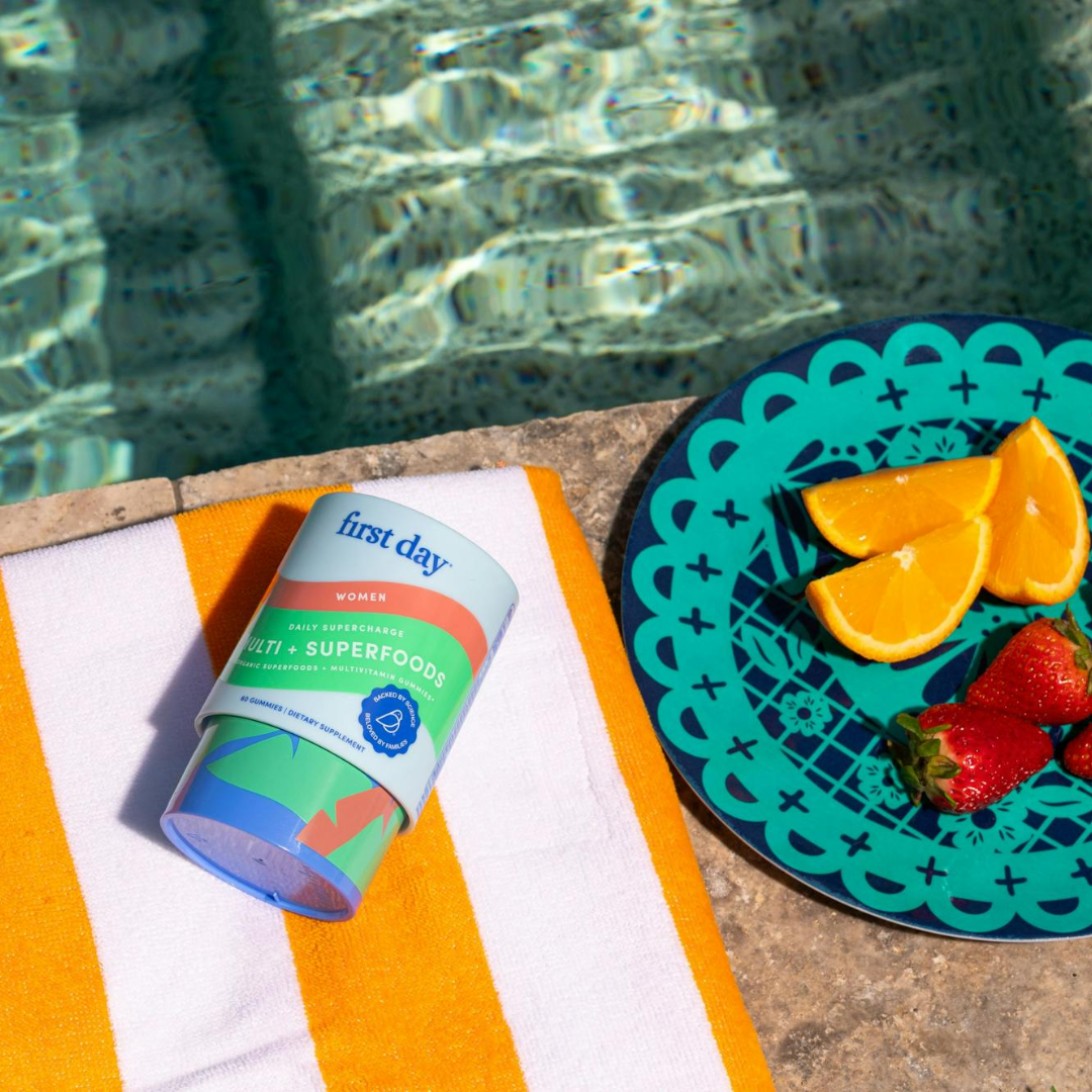 A container of First Day vitamins rests on a yellow and white striped towel beside a pool, next to a teal plate with orange slices and strawberries. Sunlight reflects off the water.