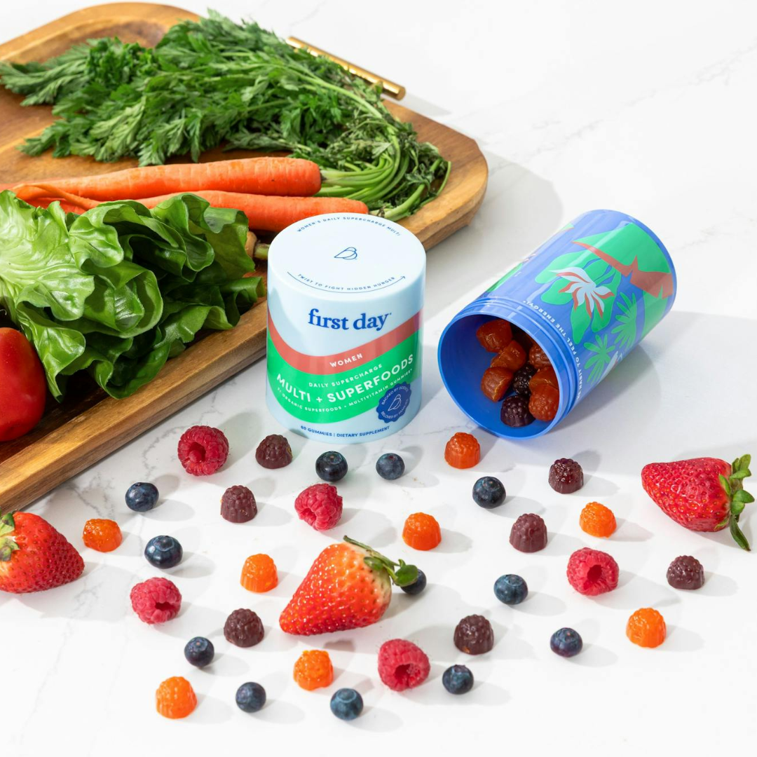 A container of First Day Women’s Multis + Superfoods gummies is on a white surface surrounded by fresh fruits, loose gummies, and an open blue tropical-themed container, with leafy greens and carrots on a wooden tray.